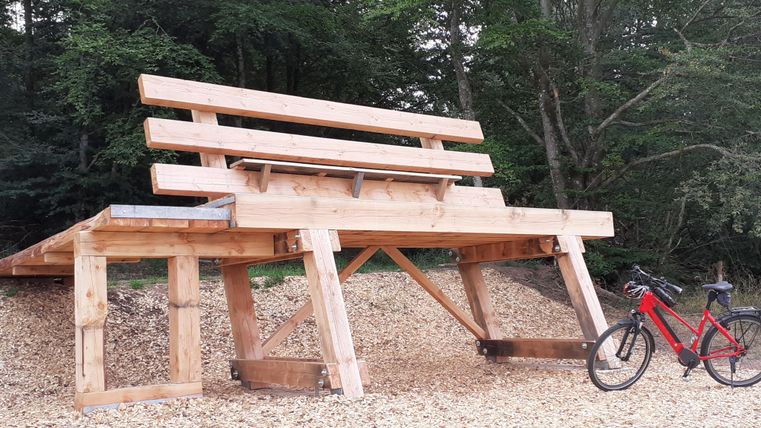 An oversized wooden bench in the forest, where a person is sitting. Next to it is a red bicycle. The ground is covered with wood chips.