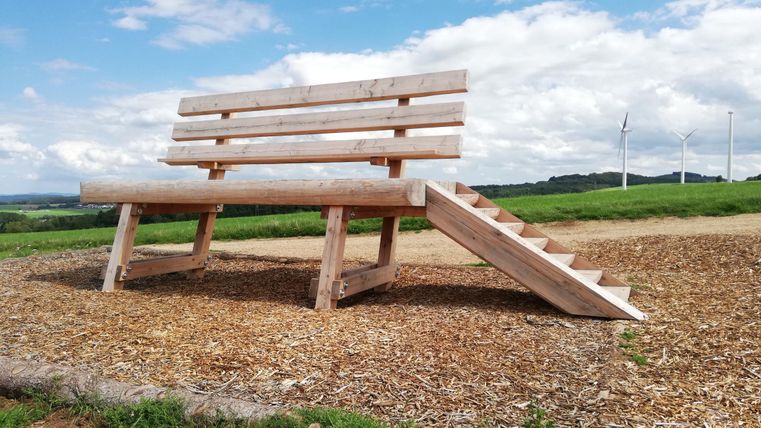 An oversized wooden bench with stairs stands in a field. In the background, wind turbines and a cloudy sky can be seen.