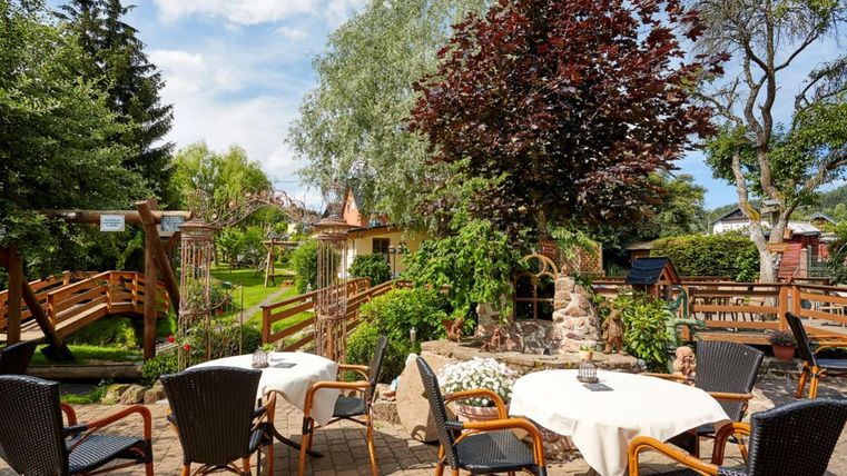 Ein einladender Außenbereich mit Tischen und Stühlen, umgeben von üppigem Grün und Bäumen. Im Hintergrund gibt es einen kleinen Holzsteg, der zu einem idyllischen Garten führt.