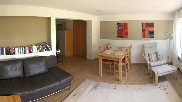 A bright and inviting living room with a black sofa, a dining table, and modern artworks on the wall. Large windows provide plenty of daylight and a pleasant atmosphere.