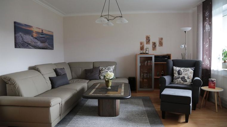 Ein gemütliches Wohnzimmer mit einer beigen Couch und einem schwarzen Sessel. Der Tisch in der Mitte ist aus Holz, und es gibt eine kleine Blumendekoration.