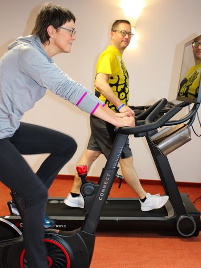 Zwei Personen trainieren in einem Fitnessraum. Eine Frau fährt auf einem Heimtrainer, während ein Mann auf einem Laufband läuft.