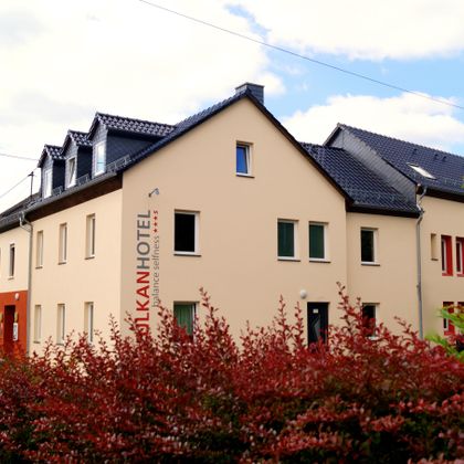 Ein modernes Hotelgebäude mit einem klaren, freundlichen Design. Im Vordergrund sind rotbunte Büsche zu sehen, die eine einladende Atmosphäre schaffen.