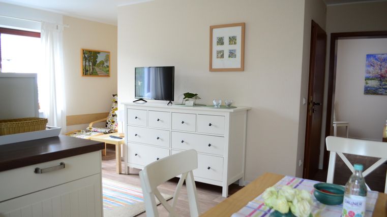 A bright, modern living room with a television and a white dresser. The room features an inviting dining table with flowers and a water pitcher.