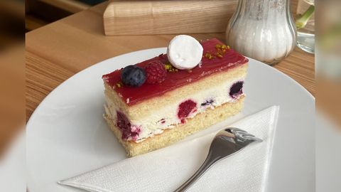 Een stuk slagroomtaart met een fruitige laag van frambozen en bosbessen. De taart ligt op een wit bord met een lepel en een servet.