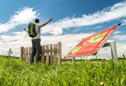 Een wandelaar staat met uitgestrekte armen voor een houten hek op het Vulcano Trail. Op de voorgrond is een informatiebord zichtbaar.