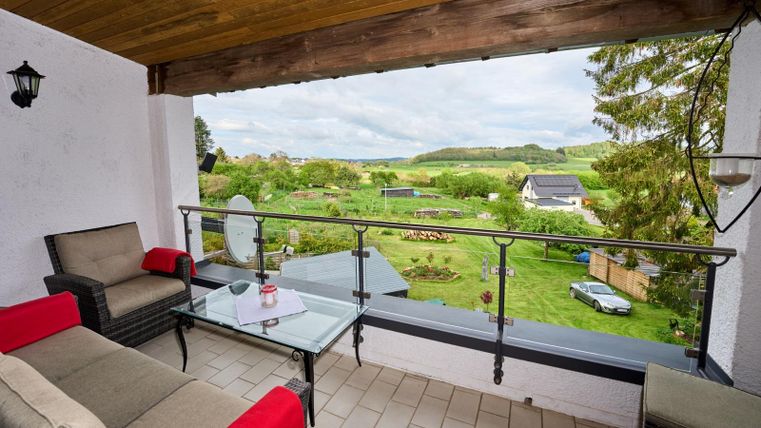 Een gezellige terrace met een glazen tafel en comfortabele meubels. Het uitzicht gaat naar een groene landschap met tuinen en heuvels op de achtergrond.