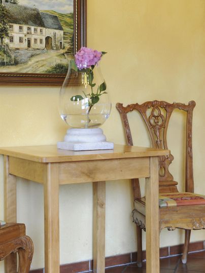 Ein Holztisch mit einer Blumenvase und einer einzelnen Blume steht in einem hellen Raum. Im Hintergrund hängt ein Bild eines Hauses.