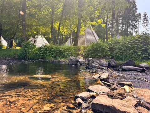 Ein ruhiger Fluss mit klarem Wasser und Steinen im Vordergrund. Im Hintergrund sind Zelte und üppige Bäume zu sehen.