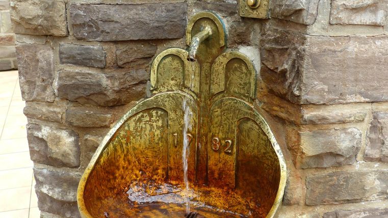 Ein rustikaler Trinkbrunnen aus Metall mit fließendem Wasser. Die Umrandung ist aus Stein und zeigt alte, unregelmäßige Strukturen.