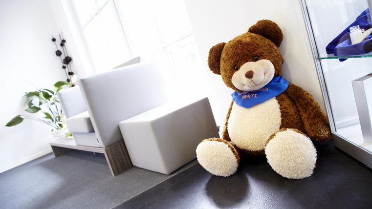 Een grote, zachte teddybeer zit in een lichte kamer. Daarnaast zijn er moderne meubels en een paar planten.