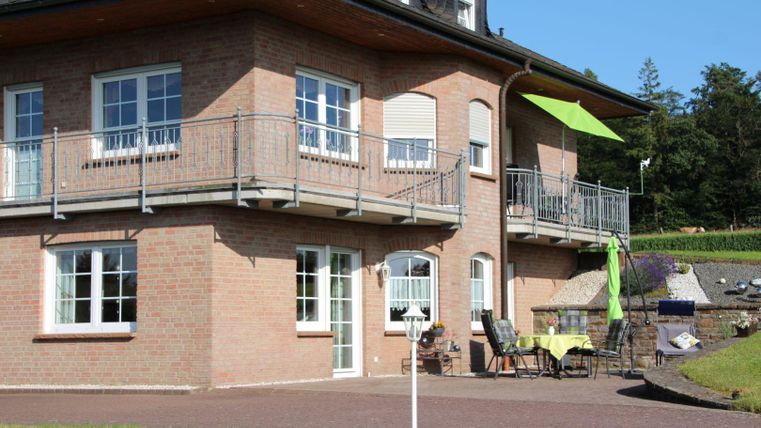 Ein modernes Haus mit einem großen Balkon und einem grünen Sonnenschirm. Vor dem Haus befinden sich Gartenmöbel und eine gepflegte Rasenfläche.