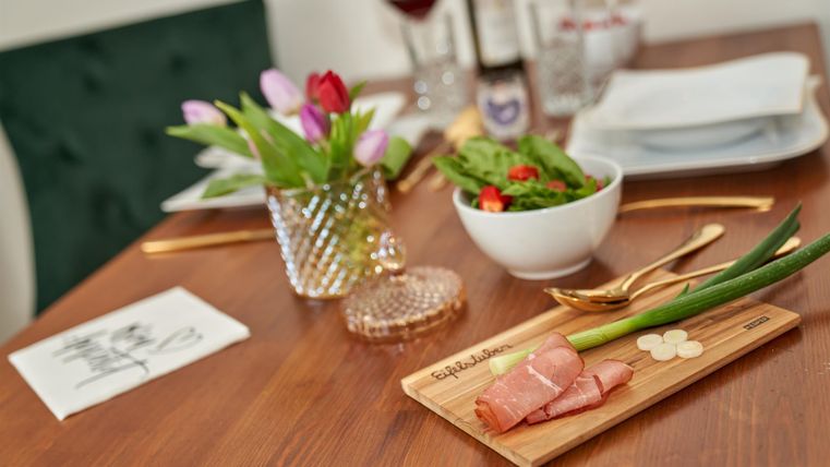 A beautifully set table with fresh ingredients. It features plates, a salad bowl, a flower vase, and elegantly arranged ham.