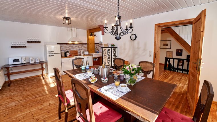 A cozy dining area with a large wooden table and chairs. The kitchen in the background is bright and inviting.