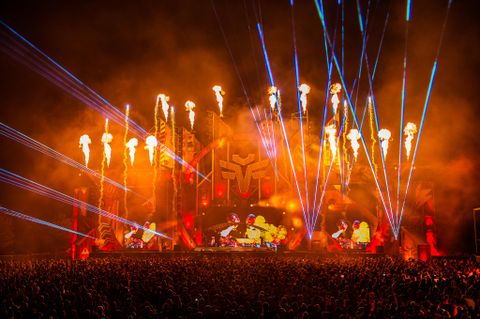 Een levendig muziekfestival met een grote menigte. Kleurrijke lichtstralen en vuurwerk verlichten het podium.