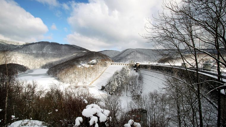 A winter landscape with snow-covered hills and a bridge. The sky is clear with a few clouds.