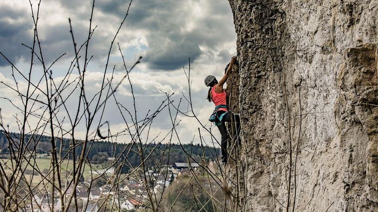 Eine Person klettert an einer steilen Felswand. Im Hintergrund ist ein Dorf und bewölkter Himmel zu sehen.