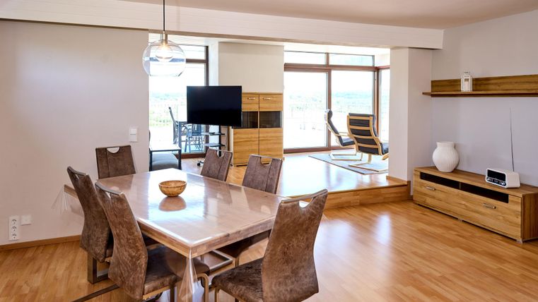 Ein modernes, helles Wohnzimmer mit einem Esstisch und stilvollen Stühlen. Große Fenster bieten viel Tageslicht und einen Blick auf die Umgebung.