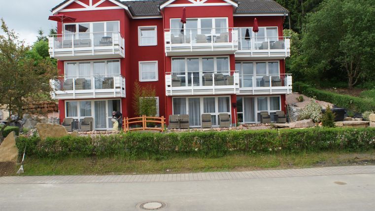 Ein rotes Wohngebäude mit mehreren Balkonen. Der Gartenbereich ist gepflegt und umgeben von Bäumen.
