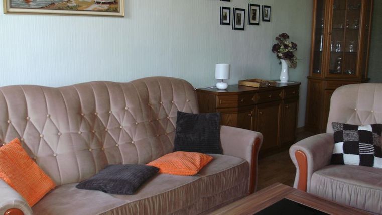 Ein gemütliches Wohnzimmer mit beigen Sofas und Dekokissen. An der Wand hängen Bilderrahmen und ein Regal aus Holz steht in der Ecke.