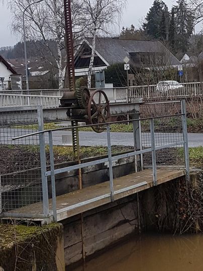 Ein Wasserregulierungsmechanismus mit einem großen Rad steht über einem flachen Gewässer. Im Hintergrund sind Bäume und ein Wohngebäude sichtbar.