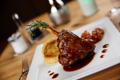 A tender lamb shank steak on a white plate, garnished with rosemary. Next to it are some cherry tomatoes and a piece of polenta.