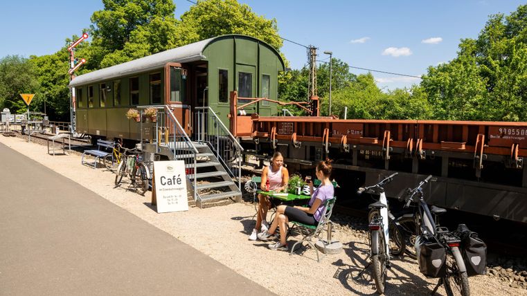 Ein Café in einem umgebauten Zugwagen, umgeben von Bäumen. Zwei Frauen sitzen draußen und genießen ihre Zeit.