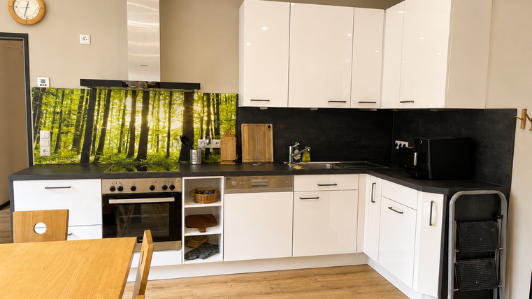 Eine moderne Küche mit weißen Schränken und einem dunklen Arbeitsbereich. Im Hintergrund befindet sich eine auffällige Wandverkleidung mit einem Waldmotiv.