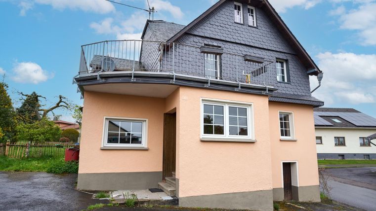 A modern house with a bright, plastered ground floor and a slate-roofed upper floor. The building has a balcony and is located in a quiet environment.