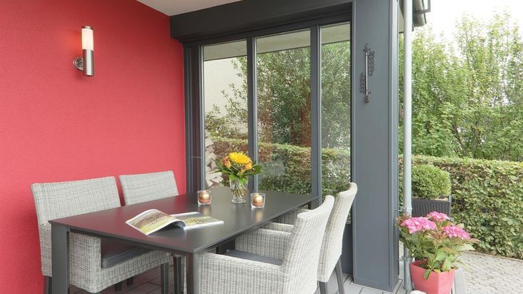 Een stijlvolle balkon met een tafel en stoelen van riet. Op de tafel staan bloemen en kaarsen, terwijl op de achtergrond groen loof zichtbaar is.