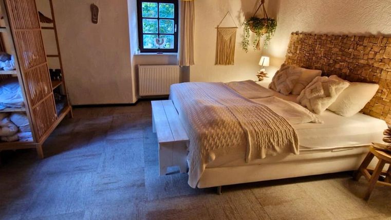 A cozy bedroom with a large bed, surrounded by warm light. The room has a natural atmosphere with wooden details and plants.