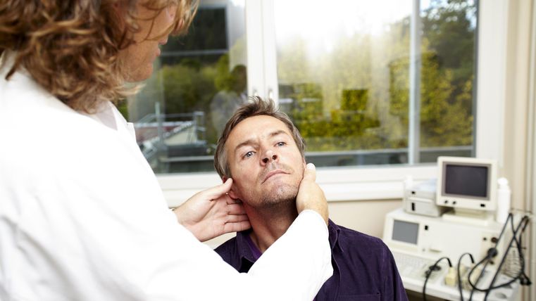 Een man zit in een dokterskamer en wordt door een professional onderzocht. Op de achtergrond zijn medische apparaten te zien.