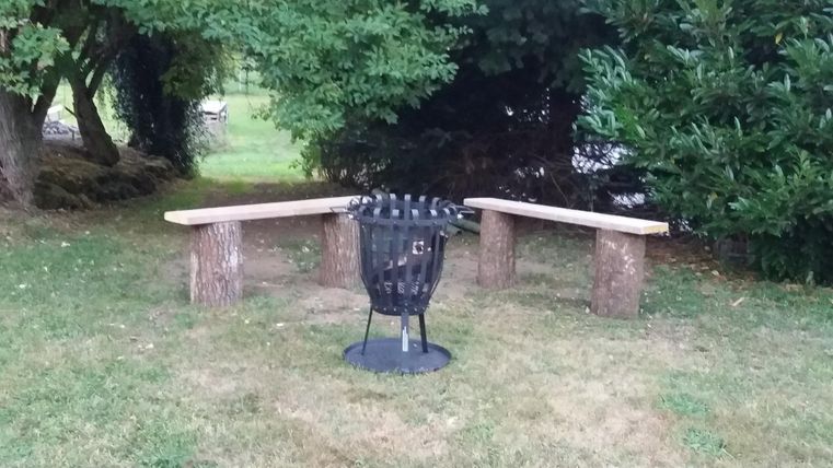 A cozy seating area outdoors with two wooden benches and a fire pit in the middle. Surrounded by green spaces and trees.