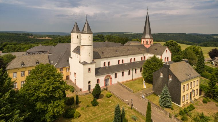 Eine historische Klosteranlage mit markanten Türmen und umgebenden Gärten. Die Landschaft ist grün und hügelig im Hintergrund.