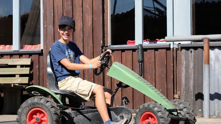 A boy is sitting on a small tractor and smiling. In the background, wooden panels and windows can be seen.