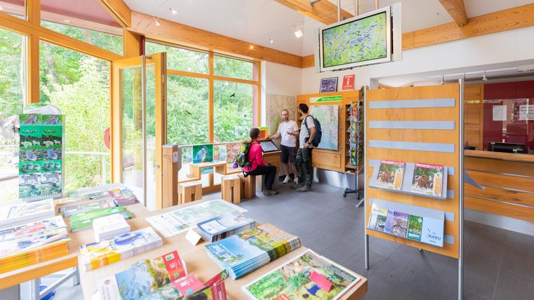 Ein Besucherzentrum mit Holzmöbeln und Informationsmaterialien. Menschen stehen an einem Empfangstresen und schauen auf eine Karte.