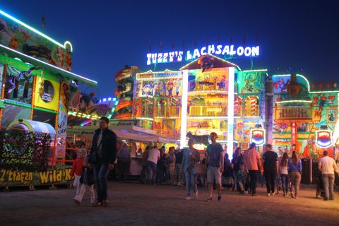 Een kleurrijke kermis met fel verlichte attracties. Veel mensen slenteren rond en genieten van de feestelijke sfeer.
