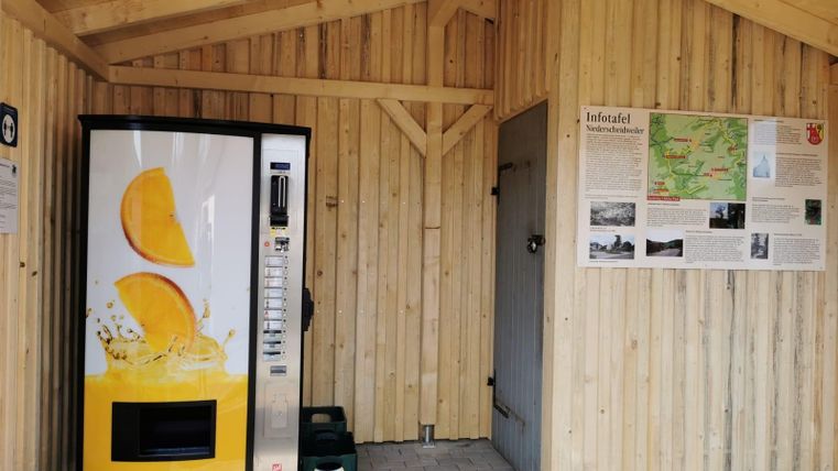 Ein Getränkewagen mit einer großen Auswahl an frischem Saft steht an einer Holzwand. Neben dem Automaten gibt es Informationen über die Region.