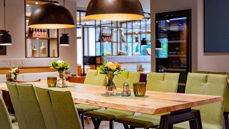 Ein eleganter Essbereich mit einem großen Holztisch und grünen Stühlen. Die Atmosphäre wird durch stilvolle Pendelleuchten und frische Blumen verschönert.
