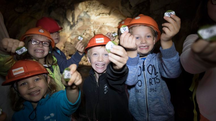 Eine Gruppe von fröhlichen Kindern trägt Helme und zeigt stolz ihre Funde. Sie stehen in einer Höhle und lächeln begeistert in die Kamera.