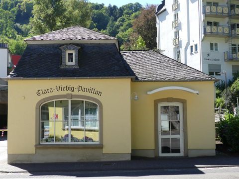 Ein kleines, gelbes Gebäude mit einem charakteristischen Dach und großen Fenstern. Über dem Eingang steht "Clara-Viebig-Pavillon".