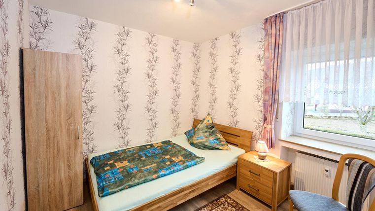 Ein einfaches Schlafzimmer mit einem Einzelbett, einem Nachttisch und einem Stuhl. Der Raum hat eine helle Tapete mit dem Muster von Pflanzen und ein Fenster, das Tageslicht hereinlässt.