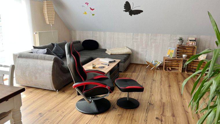 Een gezellige woonruimte met een grijze bank, een moderne stoel en een tafel. Aan de muur zijn decoratieve vlinders en planten te zien.