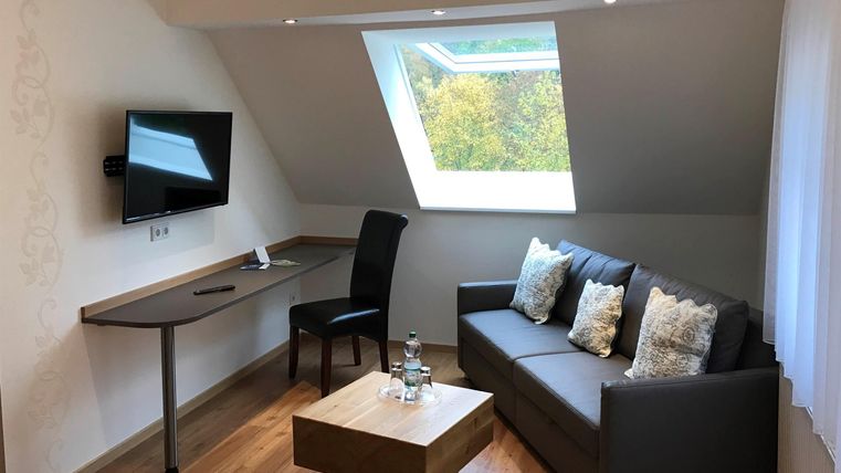 Ein gemütlicher Wohnbereich mit einem Sofa, einem Tisch und einem Fernseher. Ein Schreibtisch steht bereit, und ein Fenster bringt Tageslicht in den Raum.