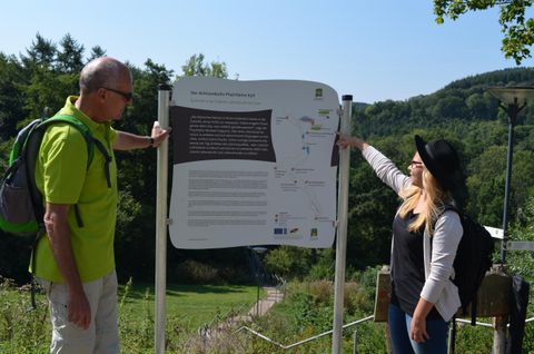Two hikers are looking at an information board in nature. In the background, trees and hills can be seen.