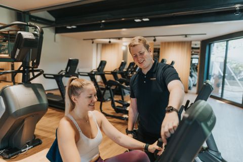 Een vrouw traint in de sportschool met een trainer. De ruimte is modern en goed verlicht, met verschillende fitnessapparaten op de achtergrond.