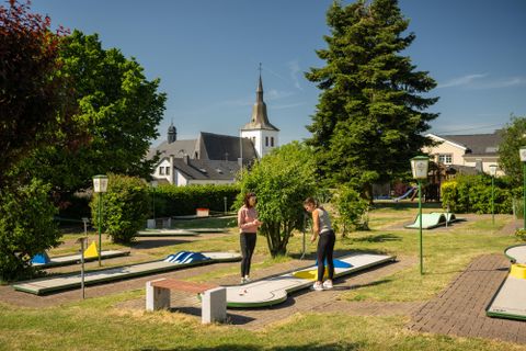 Een midgetgolfbaan met meerdere banen en groenvoorzieningen. Op de achtergrond is een kerk en blauwe lucht te zien.