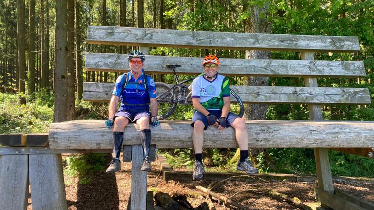 Two cyclists are sitting on a large wooden bench in the woods. Trees are visible in the background, and the sun is shining.