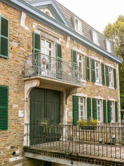 Een historisch gebouw met een stenen gevel en groene luiken. De ingang omvat een balkon met een smeedijzeren balustrade.