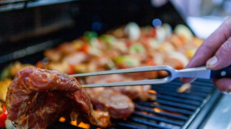 Eine Hand hält ein Stück gegrilltes Fleisch auf einem Grill. Im Hintergrund sind Spieße mit Gemüse und Fleisch zu sehen.